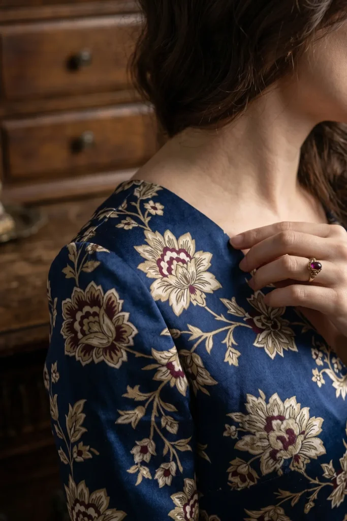 A close-up, high-detail fashion photograph focusing on the shoulder and neckline of a woman wearing a luxurious, dark navy blue silk garment. The fabric features an intricate baroque floral pattern with large cream-colored blossoms, burgundy accents, and delicate gold filigree vines. The woman has wavy brown hair draped over her neck, and her hand rests gently near her collarbone, showcasing a vintage gold ring set with a prominent oval-cut ruby gemstone. In the softly blurred background, a rich, dark-toned wooden vintage chest of drawers provides a moody, classic aesthetic.