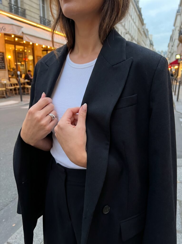 A stylish close-up shot of a woman's hands holding the lapels of a tailored, oversized black blazer that is draped elegantly over her shoulders. Underneath, a hint of a white ribbed tank top is visible. The shot captures a high-fashion, effortless European evening look.