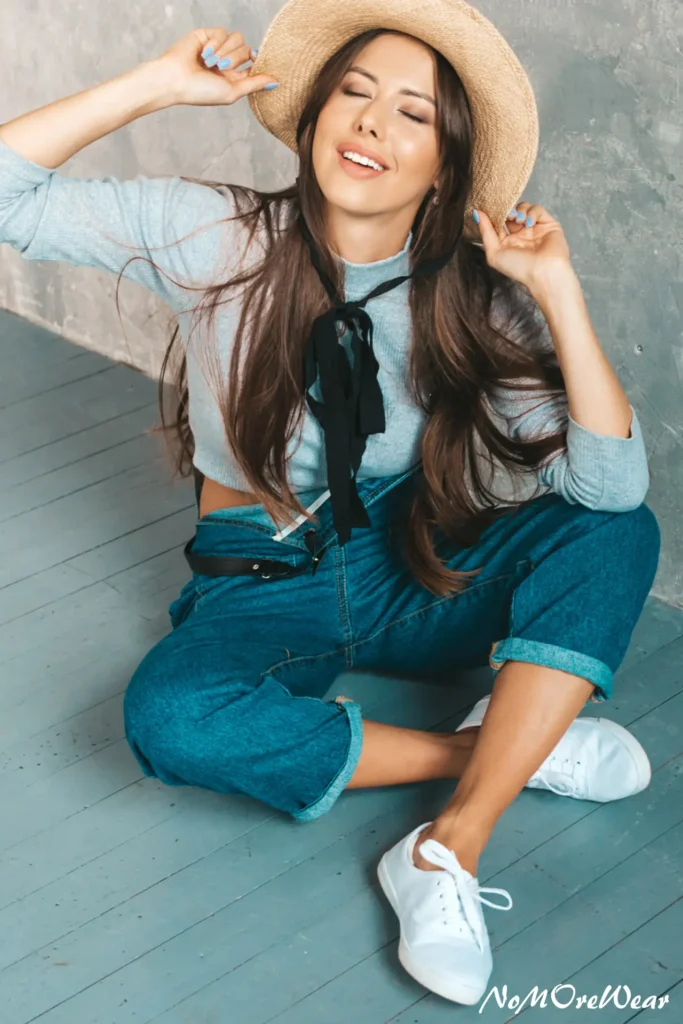 A woman sitting and smiling in a modern double denim look featuring blue denim overalls paired with a light blue long-sleeve knit top, a straw hat, and white sneakers for a vintage-inspired casual everyday outfit.