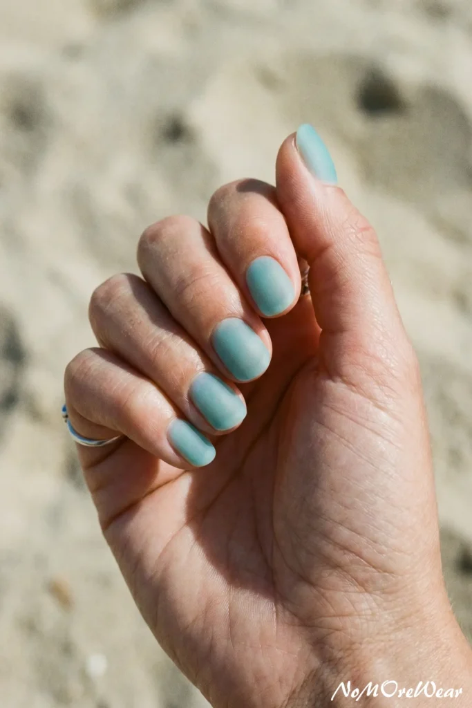 A professional close-up macro photograph focusing on a well-manicured female hand resting naturally against a soft, out-of-focus background of bright neutral sand at a beach. The short, neatly shaped square-oval nails are coated with a stunning muted aqua teal gel polish featuring a smooth frosted matte finish that mimics frosted sea glass. A simple, thin silver band ring is visible on her pinky finger. The direct natural sunlight perfectly highlights the velvety texture of the nail coating, neat cuticles, and realistic skin texture of the hand. In the bottom right corner, a small, elegant white cursive watermark reads "NoMoreWear". The entire look embodies a crisp, "clean girl" summer beauty mood, ideal for minimal nail design inspiration, beach-ready gel colors, and creative modern manicures.