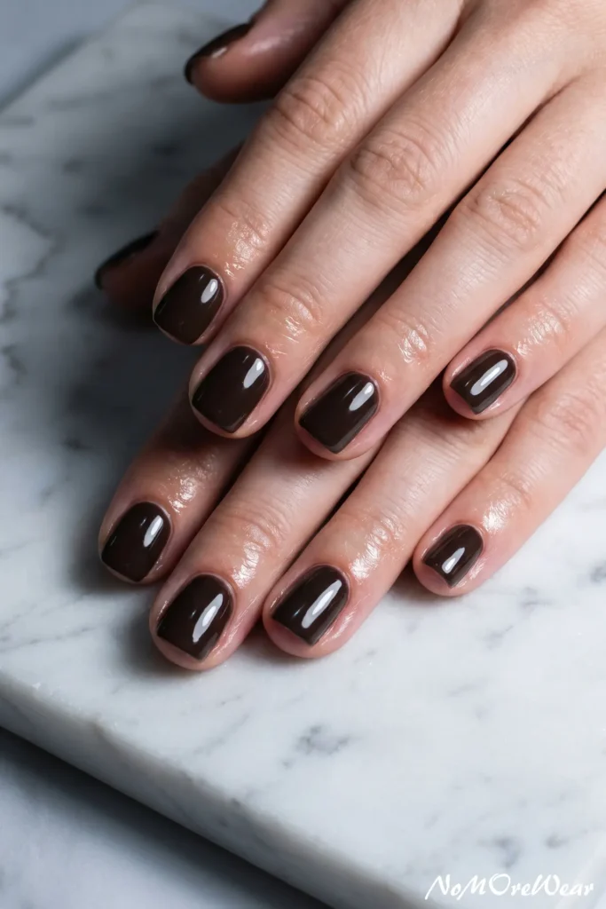 A professional close-up macro photograph showing a woman's well-manicured hands elegantly stacked together, resting flat on a polished white marble surface with soft grey veining. Her short, square-oval nails are painted in a high-gloss, ultra-deep espresso brown nail polish that delivers a rich, solid opaque finish. The dark chocolate, near-black hue shines beautifully under the studio lighting, reflecting clean light lines and highlighting the immaculate cuticles and smooth, hydrated skin texture of her fingers. In the lower right corner, a subtle, elegant white cursive watermark reads "NoMoreWear". The entire look embodies a bold "quiet luxury" and sophisticated dark winter aesthetic, perfect for everyday minimal manicure inspiration, elegant gel polish trends, and clean beauty capsule images.