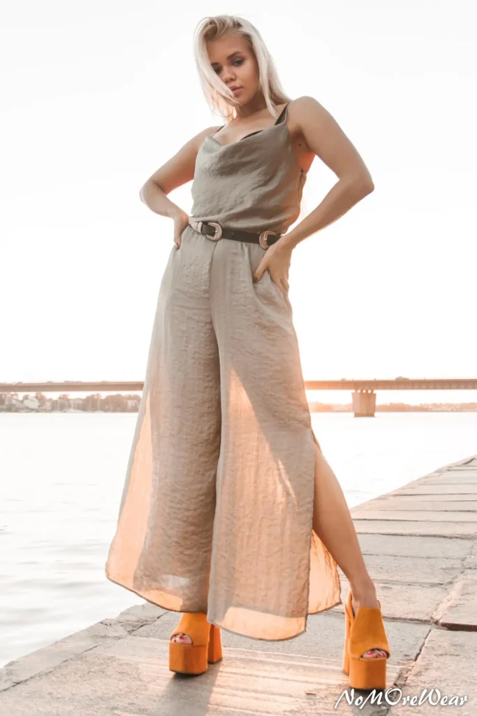 A woman standing by the water at sunset wearing elegant summer workwear: stone-colored wide-leg linen trousers with side slits, a matching cowl-neck top, and a black belt. She completes the look with vibrant orange platform heels.
