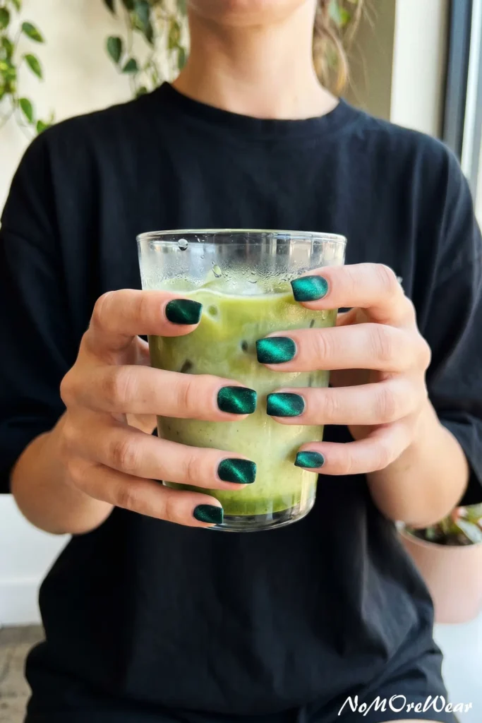 A woman holding an iced matcha latte showcasing short square emerald green velvet cat-eye nails.