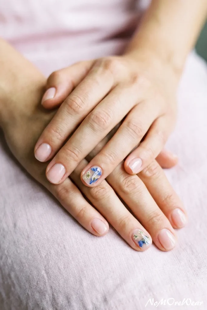 A woman's hands resting together showing a clean milky pink manicure with real dried blue flower nail art accents.