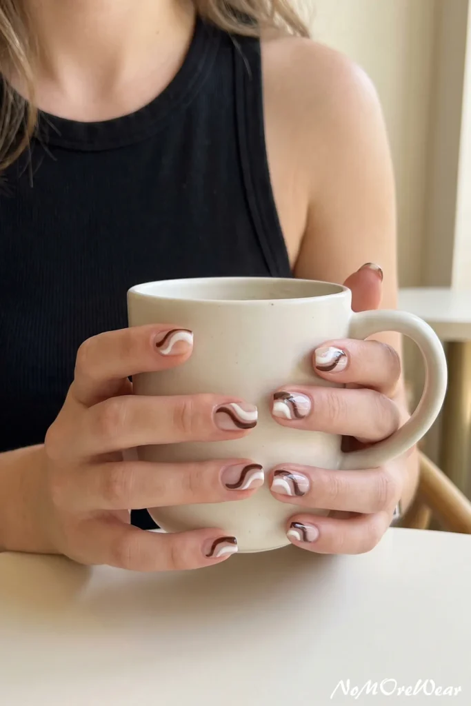 A woman holding a ceramic coffee mug showing trendy brown and white abstract swirl nail art on short square nails.