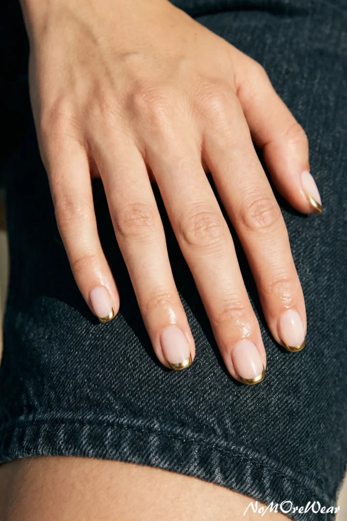 A close-up of a woman's hand resting on dark denim jeans, showcasing a minimalist French manicure with reflective metallic gold chrome tips on short oval nails.