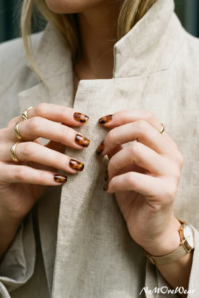 A woman adjusting her linen blazer collar, showcasing a glossy tortoiseshell nail art design on short square nails.