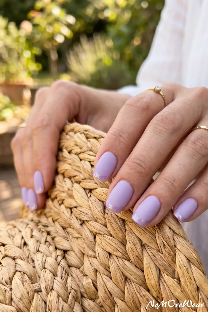 A close-up of a woman's hands resting on a woven straw tote bag, showcasing a subtle pastel lavender French manicure on short square nails.